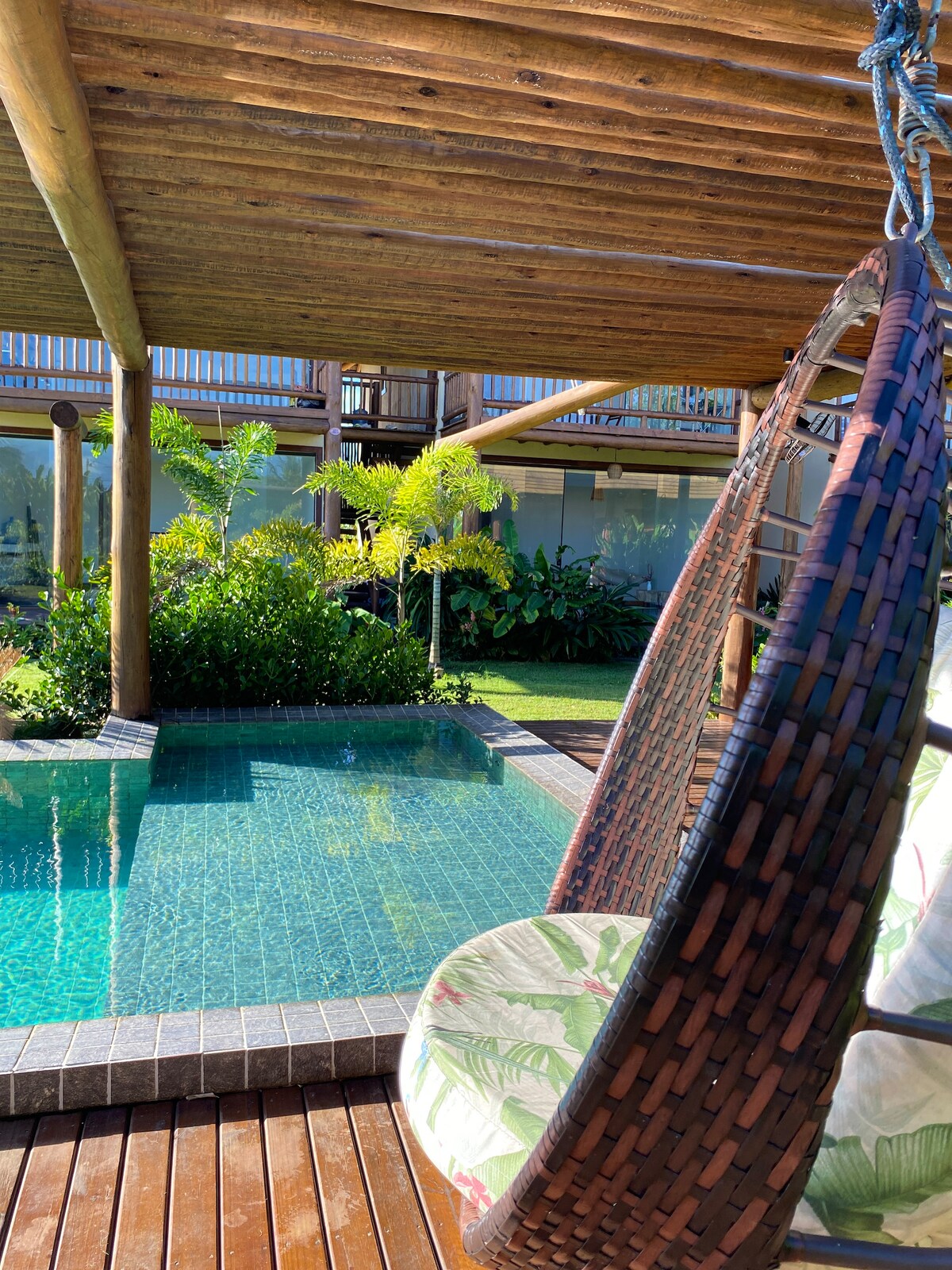 A hanging chair is positioned on a wooden deck, overlooking a tranquil pool. Lush greenery surrounds the area, complementing the natural wood structure of the overhead canopy. The pool surface reflects the sunlight, enhancing the calm ambiance.