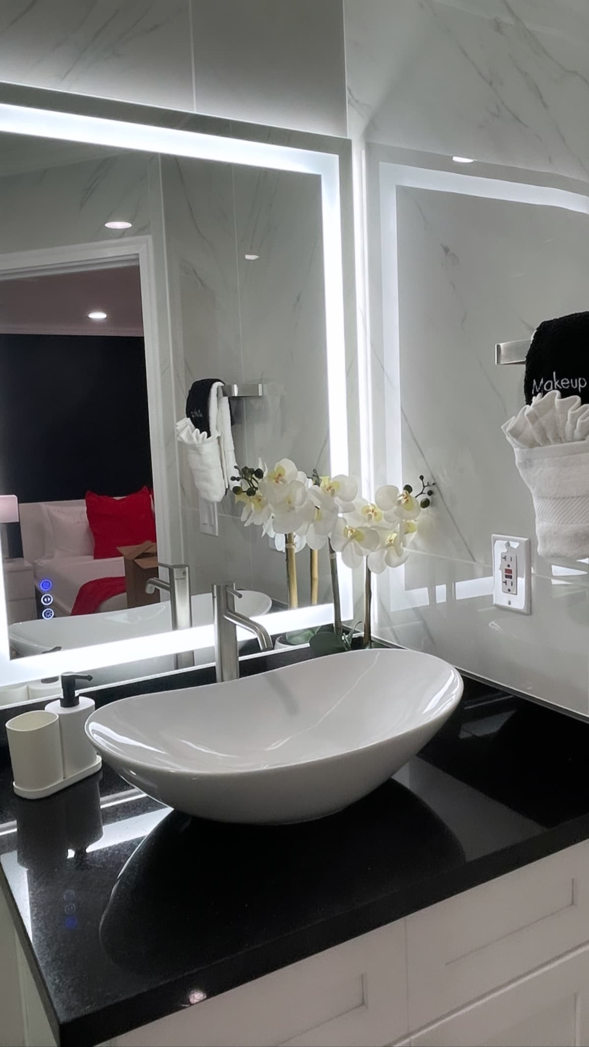 A modern bathroom is presented, featuring a sleek white vessel sink atop a dark countertop. Soft lighting highlights the contemporary design, while a decorative arrangement of artificial flowers adds a touch of elegance. The mirror and wall reflect a hint of the stylish living space beyond.