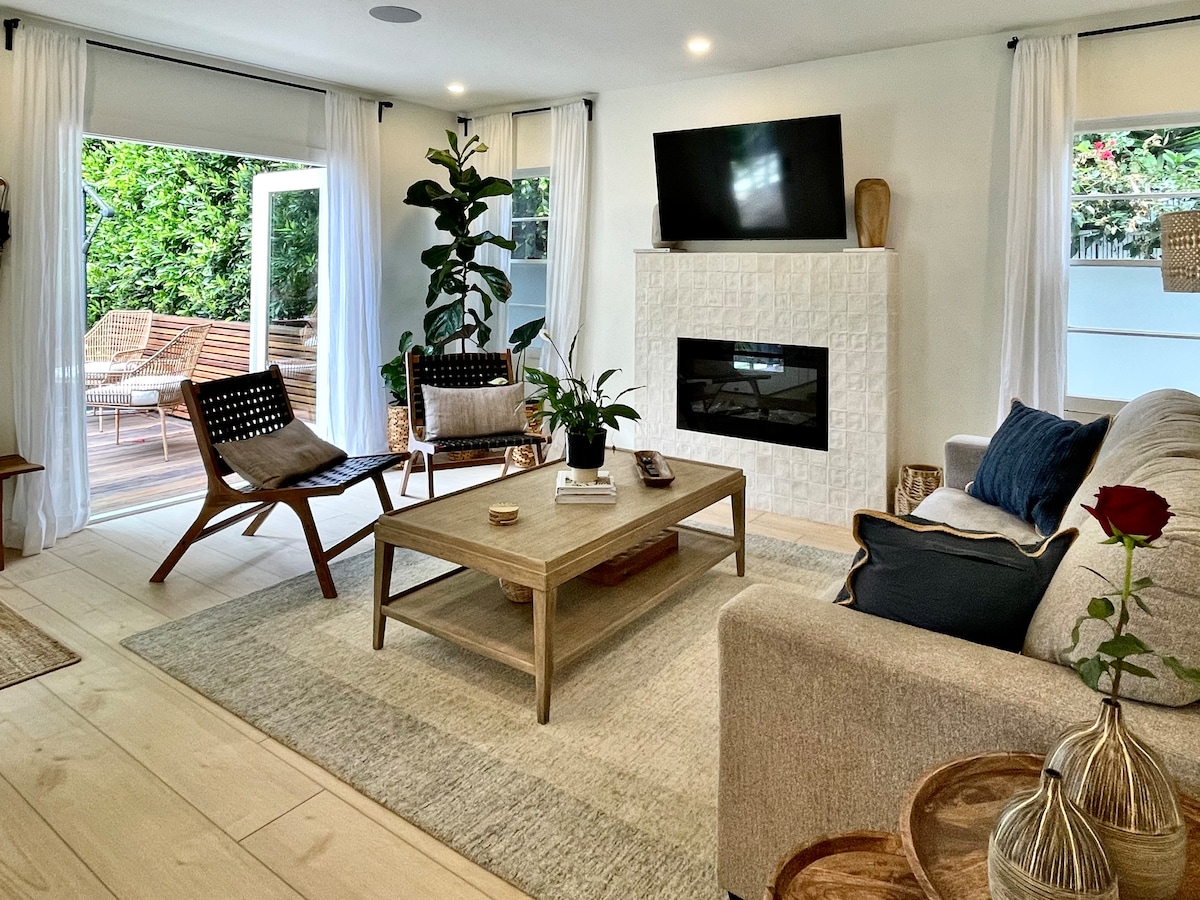 A welcoming living area features a light-colored sofa and two modern wooden chairs. A neutral rug anchors the space, while a flat-screen TV is mounted above a textured white fireplace. Large windows invite natural light, and lush greenery is visible outside.