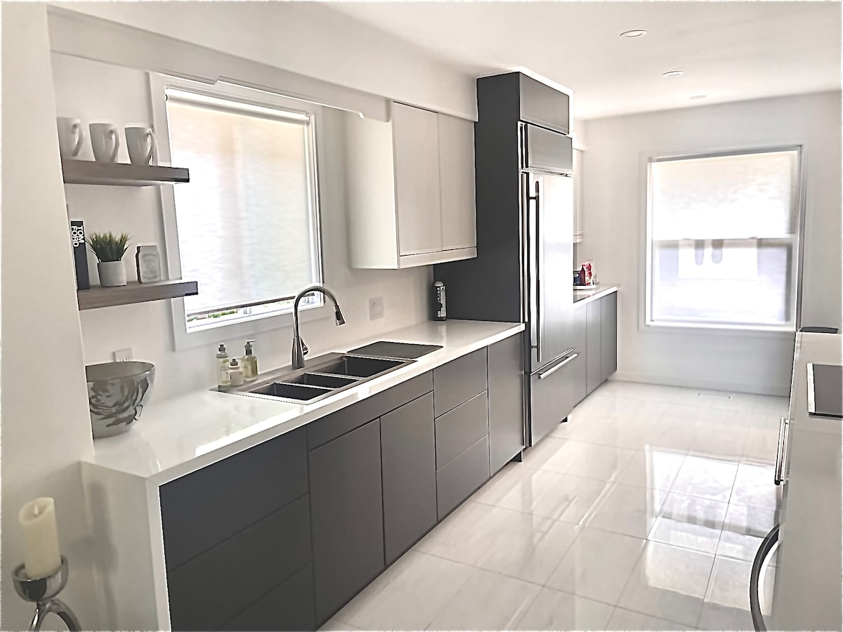 The kitchen features modern cabinetry with a mix of dark and light finishes. A double sink is set into the sleek countertop, complemented by built-in appliances and abundant natural light from the windows, enhancing the spacious atmosphere.
