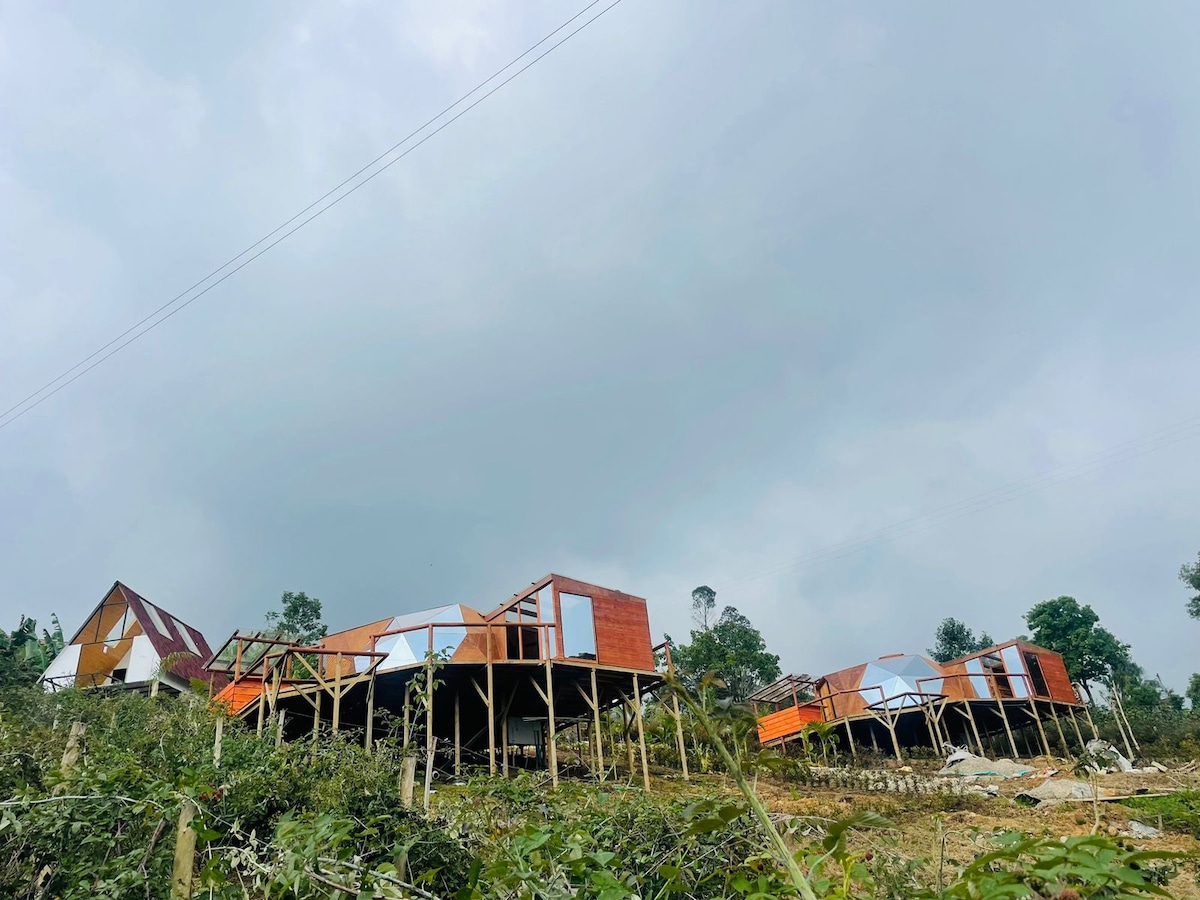 A collection of uniquely designed cabins is positioned on stilts, showcasing vibrant orange and natural wood exteriors. The structure's geometric shapes create an interesting silhouette against the cloudy sky, surrounded by a green landscape and trees in the background.