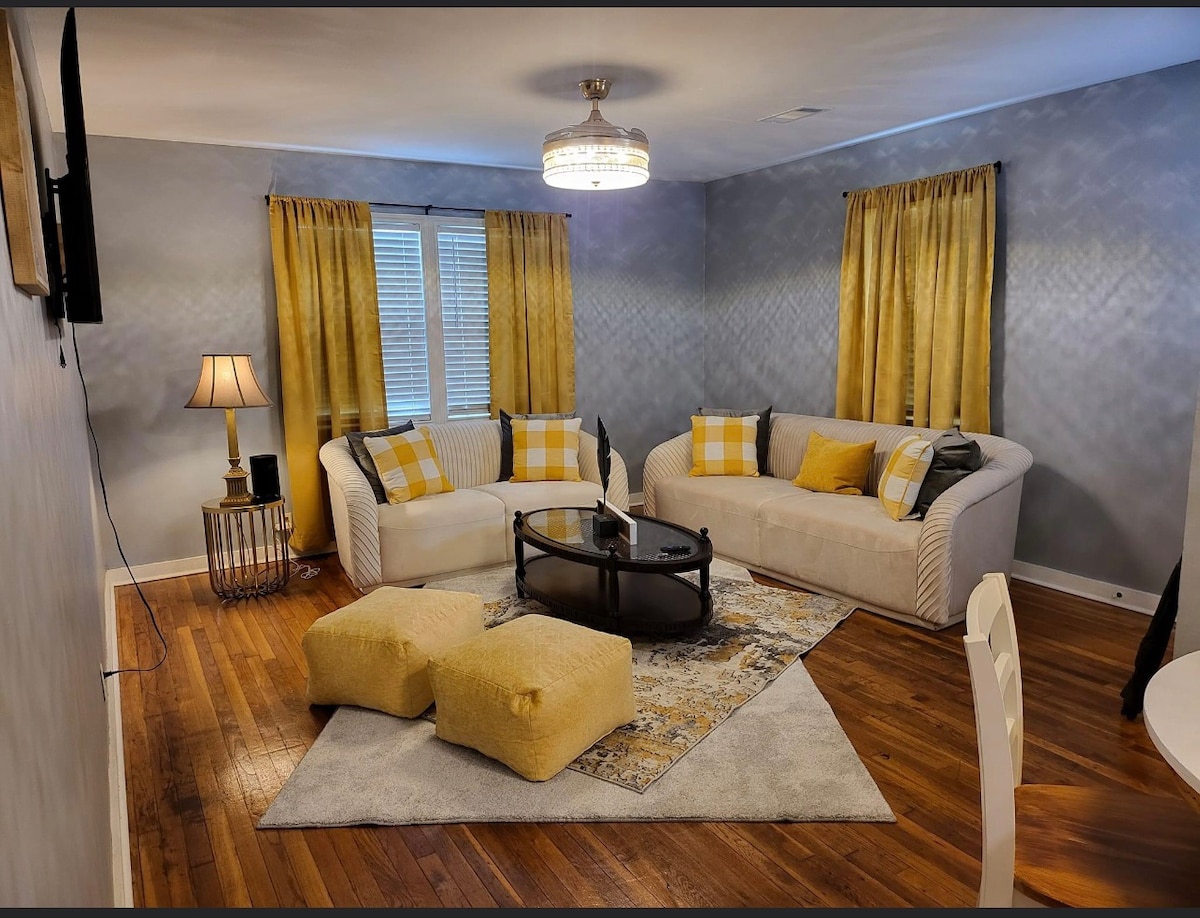 A cozy living area is presented with a white sofa and accent chairs, featuring yellow and plaid cushions. A round glass coffee table sits atop a patterned area rug. Warm lighting is provided by a stylish lamp, while yellow curtains frame the windows.