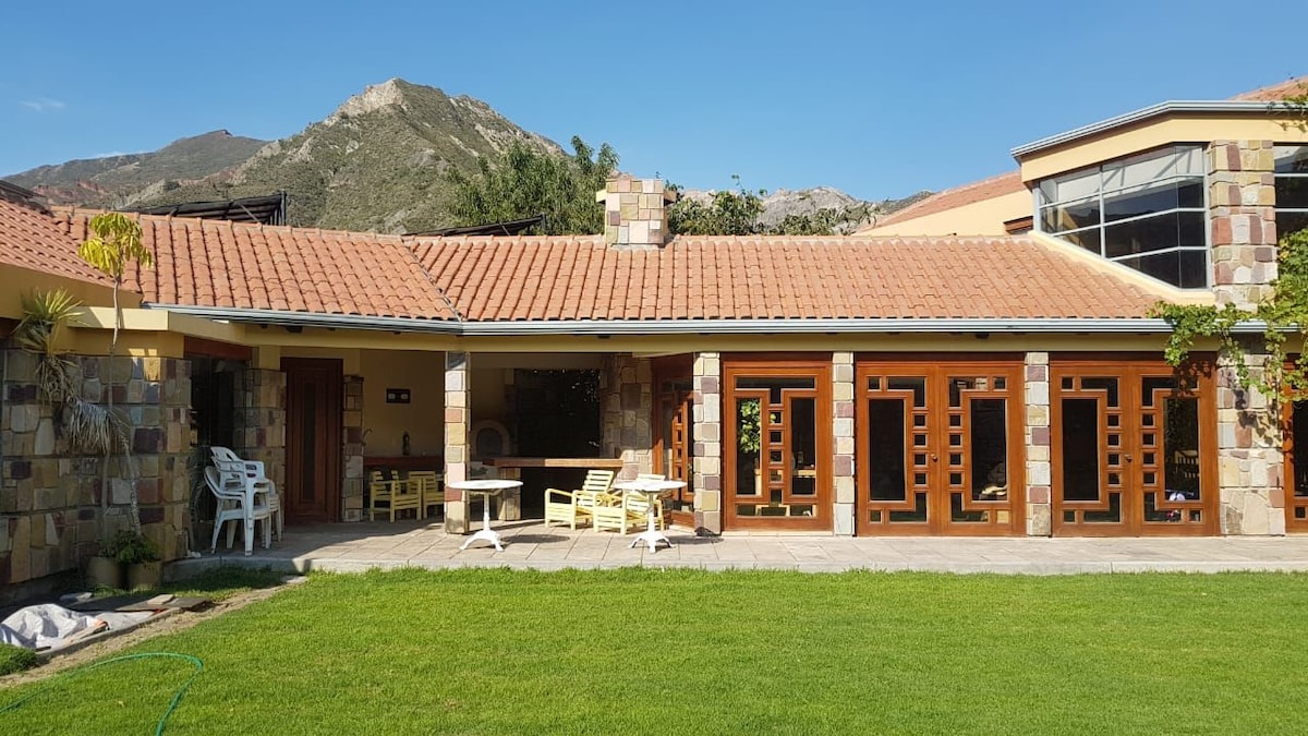 The house features a stone exterior with a red-tiled roof, complemented by large wooden doors. A spacious green lawn surrounds the structure, with outdoor seating arranged on a patio. Mountains can be seen in the background, adding to the serene atmosphere.