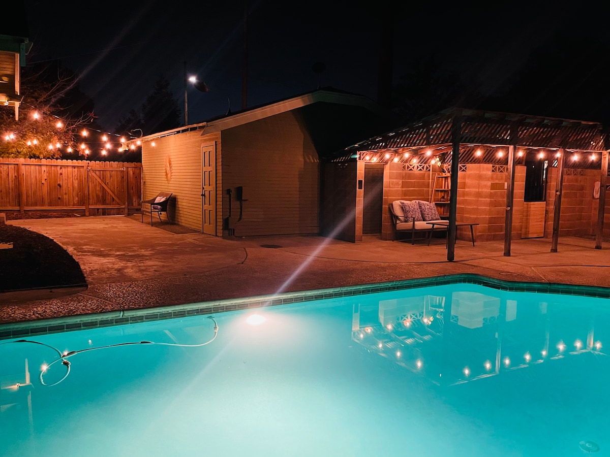 A tranquil in-ground pool glows softly under string lights at night. The pool reflects warm light, surrounded by a spacious patio area. A cozy seating area is visible nearby, along with a storage building against a fenced backdrop.