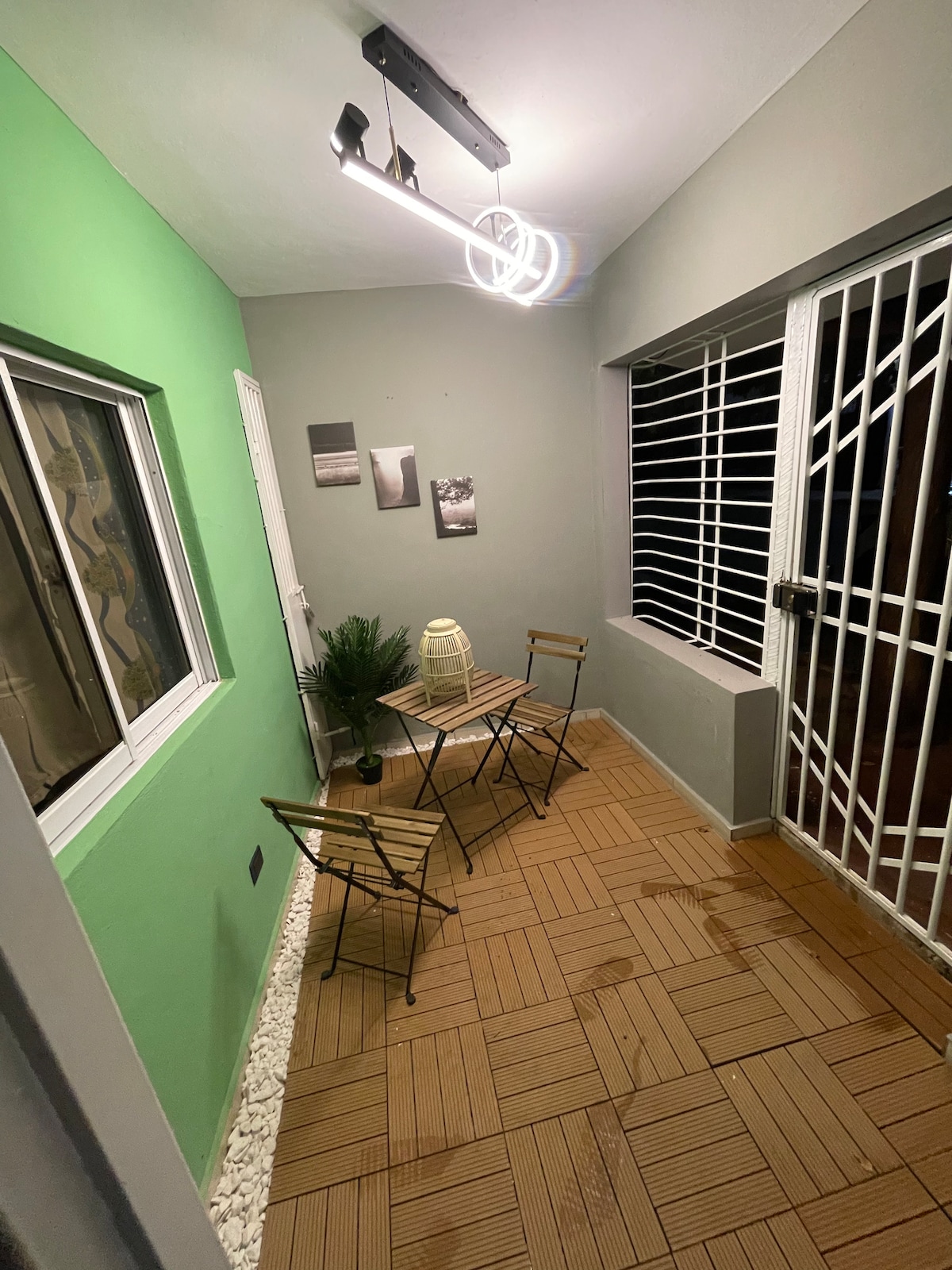 A cozy corner space features a green accent wall alongside a neutral-colored wall. Two metal chairs and a small table are positioned on a wooden floor. A potted plant and a lantern are placed on the table, while a window with grid design allows natural light to enter.