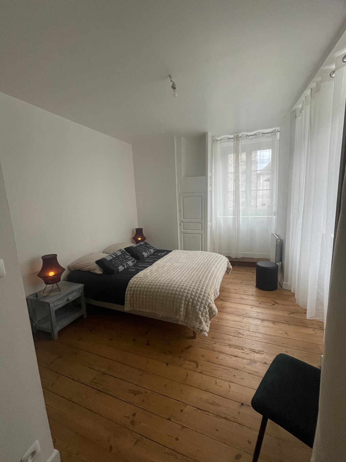 A bright bedroom features a spacious bed adorned with patterned linens. Natural light fills the room through a large window dressed in sheer curtains. Wooden floors add warmth, complemented by two bedside lamps on matching nightstands. A simple chair sits in the corner.