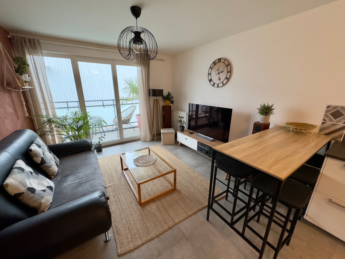 A modern living area is furnished with a black sofa adorned with patterned cushions. A wooden dining table with high chairs occupies one corner, while a flat-screen television is positioned against the wall. Large glass sliding doors provide access to a balcony, enhancing natural light.