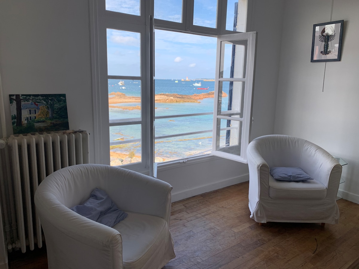 Two white armchairs are positioned by a large window, allowing a view of the sea and distant boats. Natural light floods the room, highlighting the simplicity of the decor. A small painting is displayed on the wall beside the window.
