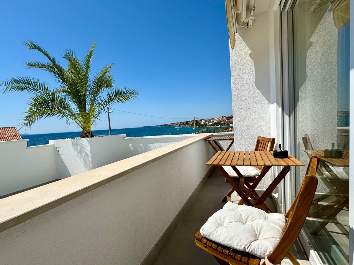 A private balcony is presented, featuring a wooden table and two chairs, inviting relaxation. A palm tree is visible, and the expansive view overlooks the beach and ocean, illuminated by bright sunlight. The vibrant blue sky enhances the serene setting.