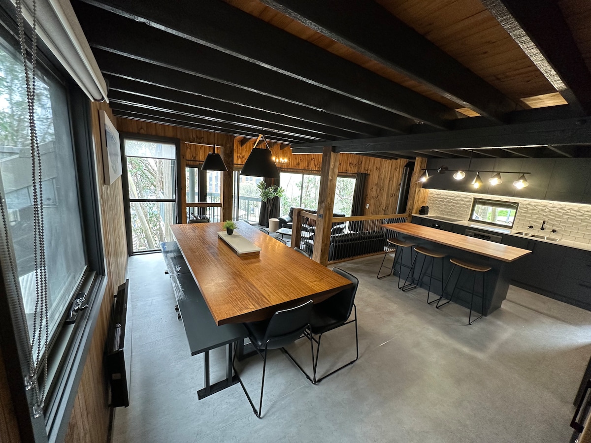A spacious dining area is complemented by a long wooden table surrounded by black chairs. An open design allows natural light to enter through large windows, showcasing black cabinetry and modern lighting fixtures in the adjacent kitchen.