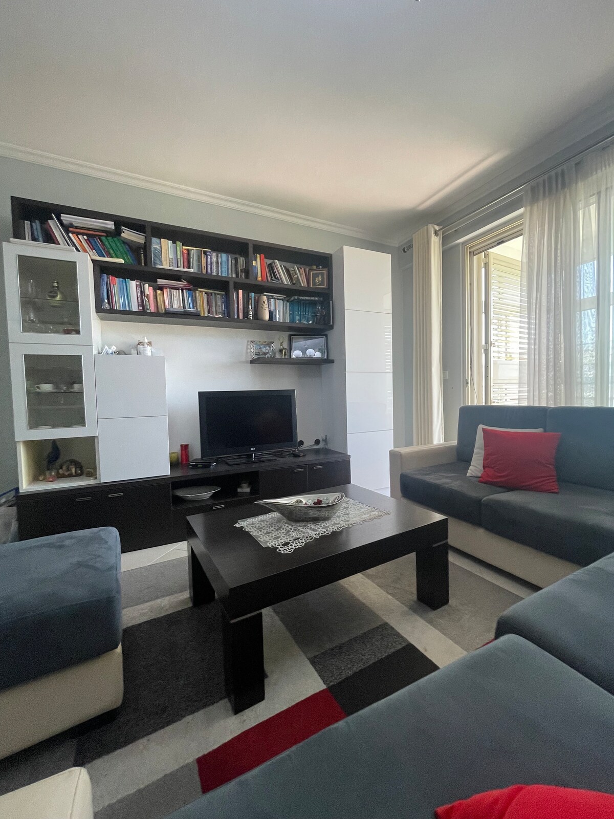 A modern living area features a low coffee table at the center, surrounded by a sectional sofa and armchairs. A large bookshelf holds various books, and a television is positioned against the wall. Natural light enters through large windows draped with sheer curtains.