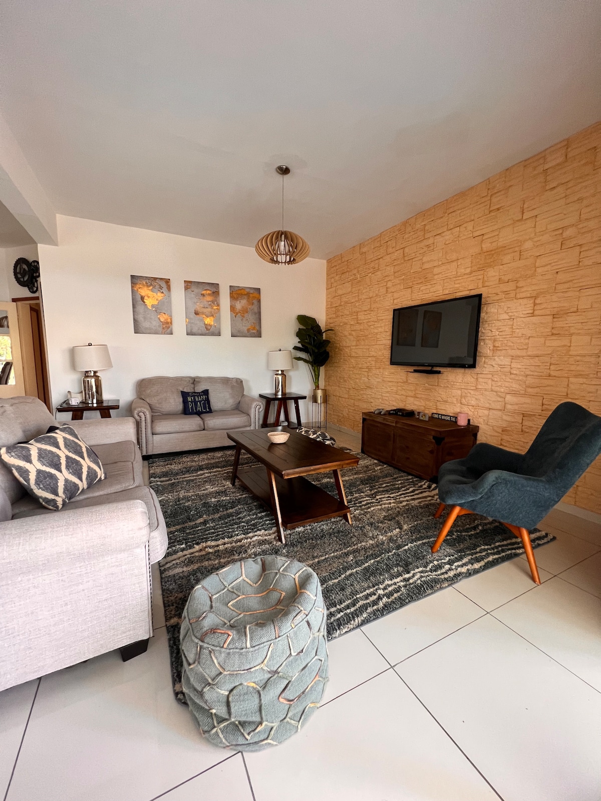 An inviting living area features comfortable seating, including a light grey sofa and an upholstered armchair in dark green. A wall-mounted television is positioned above a wooden media console. Textured artwork adorns the wall, while a patterned rug anchors the space, enhancing its cozy ambiance.