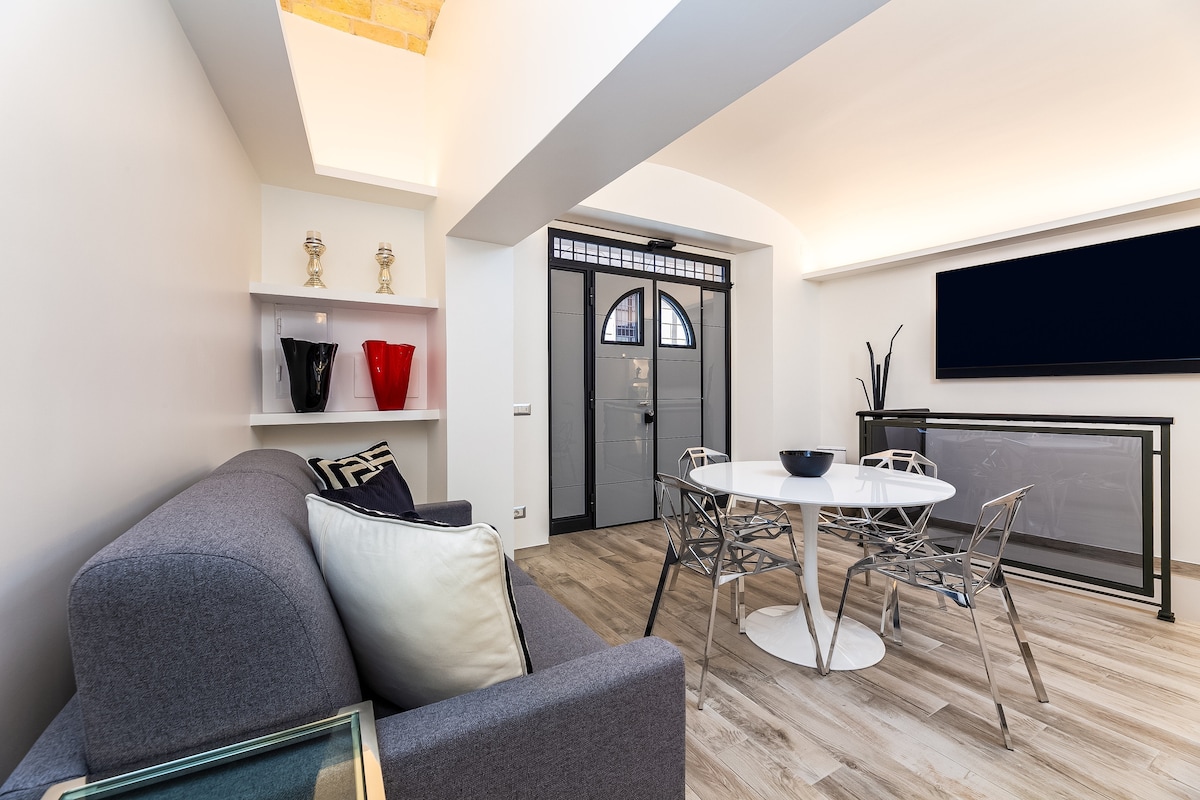 A modern living area features a comfortable grey sofa, accented with decorative cushions. A round dining table with transparent chairs is positioned nearby, complemented by a sleek TV unit. Natural light illuminates the space, highlighted by elegant decor on the shelving units.