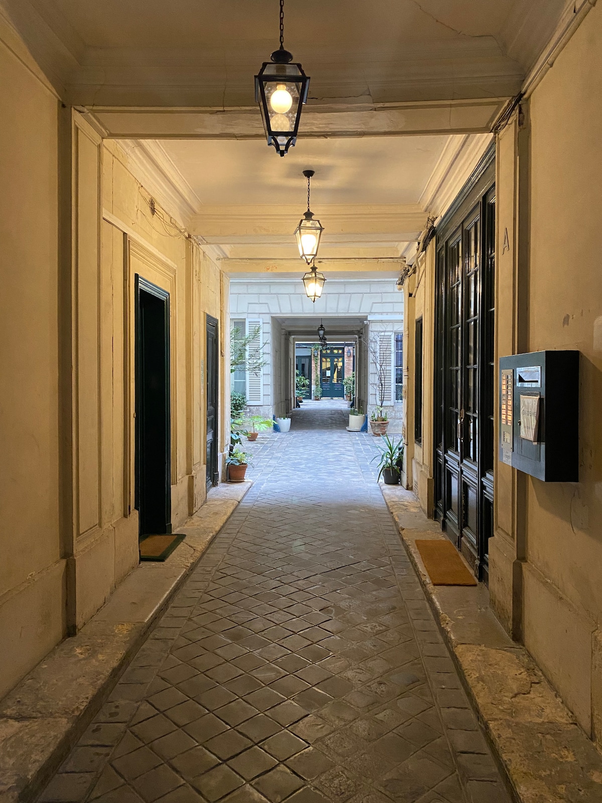 A wide corridor is illuminated by decorative lanterns hanging from the ceiling. Potted plants line the sides of the path, which is paved with rectangular stones. Doors lead to different rooms, with an open view of the courtyard at the end of the hallway.