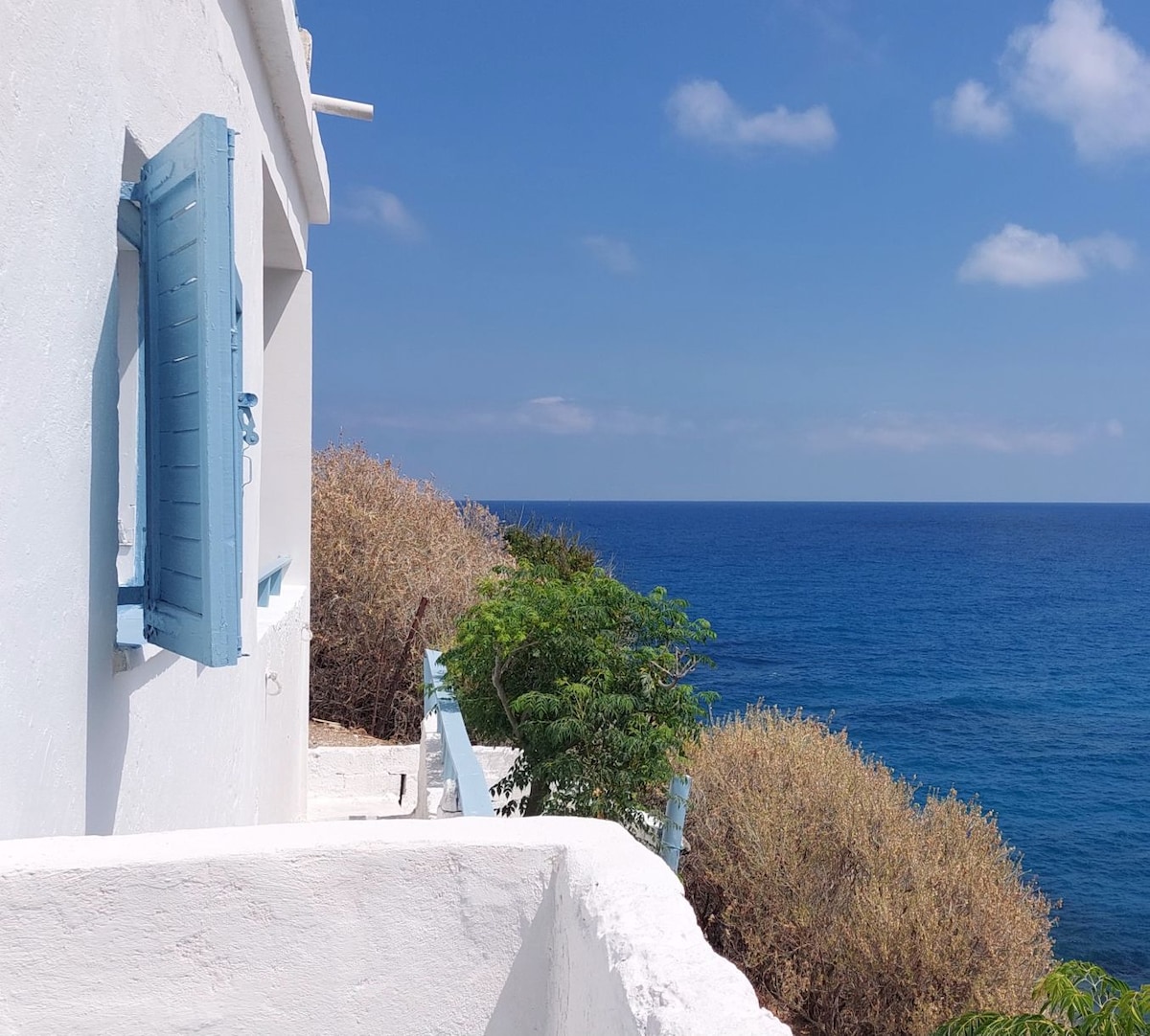 A whitewashed wall of the house is visible, featuring a blue shuttered window. The scene overlooks the deep blue sea, with greenery in the foreground adding to the coastal view. Soft clouds float in the bright blue sky.