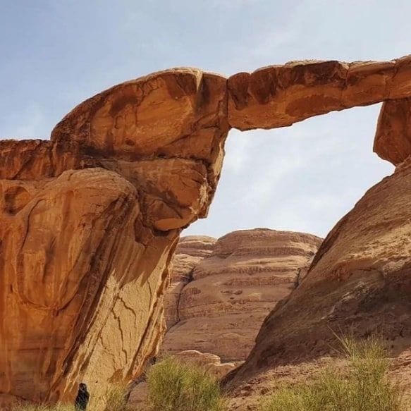 A natural rock formation resembling an arch is situated among stunning desert cliffs, showcasing layers of vibrant orange and tan stone. An individual is positioned near the base, providing a sense of scale to the impressive height and width of the arch.