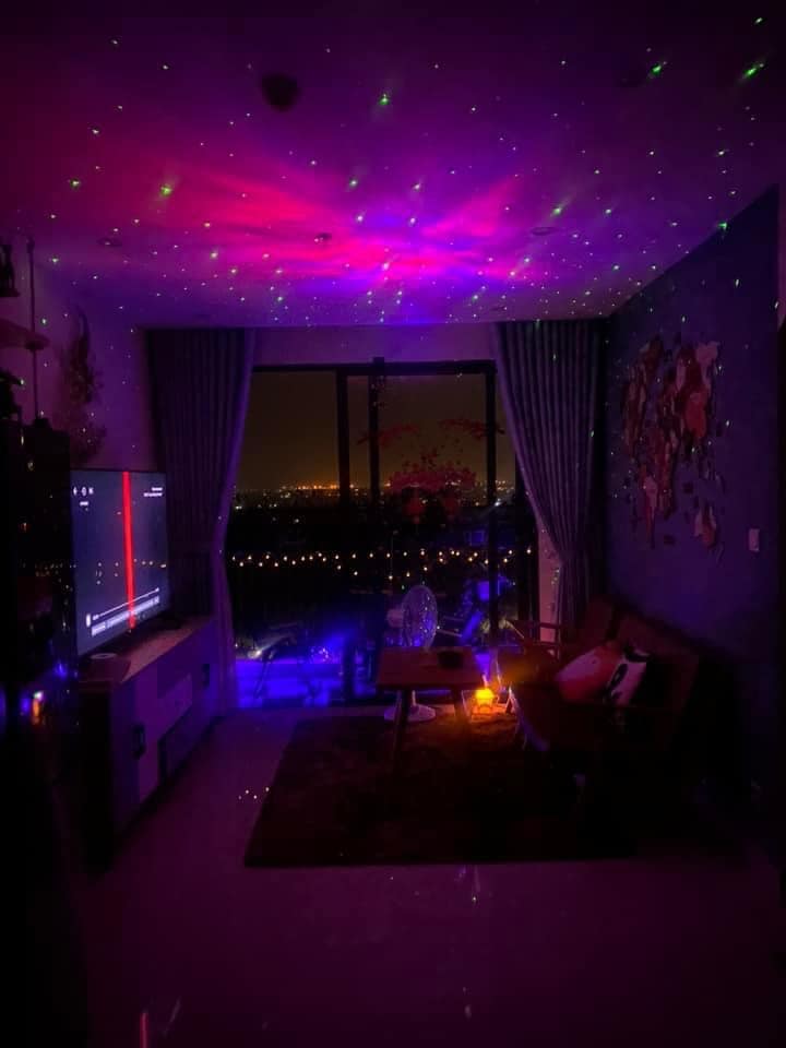 A cozy living space is illuminated by colorful lights reflecting off the ceiling. A comfortable sofa sits opposite a large television, while a small table adorned with a decorative piece and a glowing lamp creates a welcoming ambiance. Large windows offer a view of the city skyline.