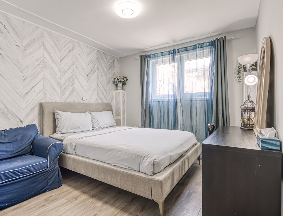 A cozy private room features a queen bed with light bedding against a pale herringbone-patterned wall. Natural light enters through sheer blue curtains at the window, illuminating a dark dresser. A blue chair and a decorative birdcage add charm to the space.
