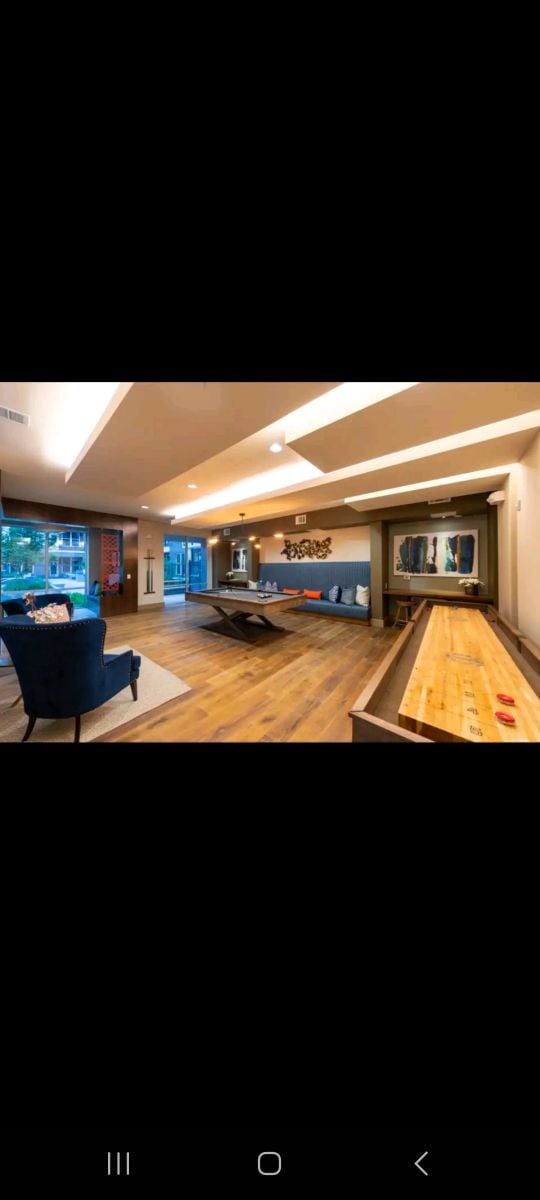 A modern living area features a wooden floor and ample natural light from large windows. A blue armchair with decorative pillows is positioned near a game table. The wall is adorned with artwork, and a sleek entertainment unit is visible.