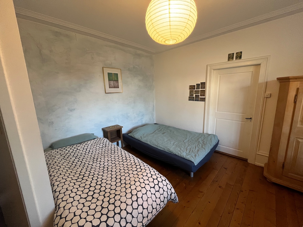 The sleeping area features two beds with light-colored bedding, arranged against a softly hued wall. A small side table is positioned next to one bed. Wooden flooring is visible beneath the beds, and a closet is present in the corner. A pendant light hangs above, illuminating the space.
