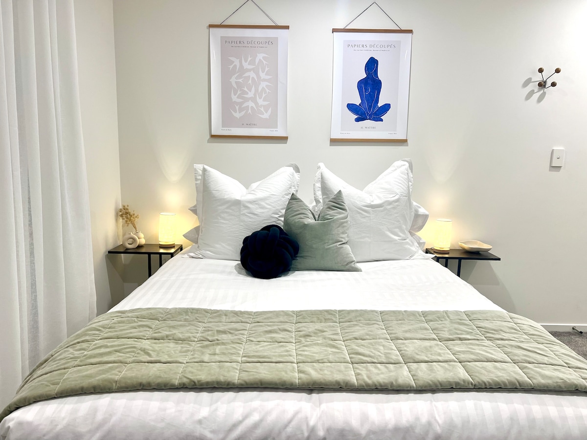 A modern bedroom features a queen bed dressed with soft white linens and an olive green throw blanket. Two decorative prints hang above the bed, complemented by bedside lamps that provide gentle illumination. Plush accent pillows add comfort and style.