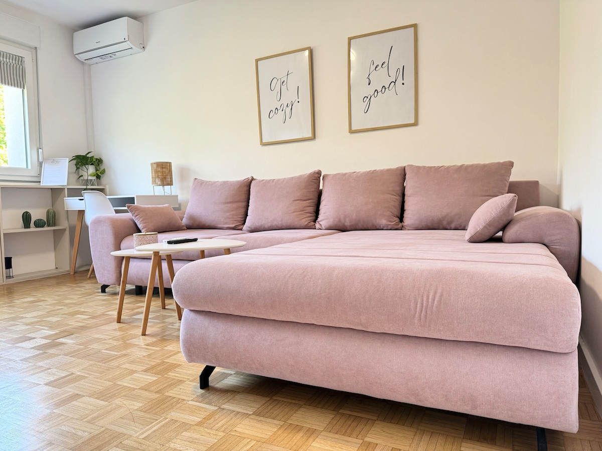 A spacious living area features a large, comfortable sectional sofa in a soft pink hue. Light wooden flooring complements the neutral decor, while air conditioning is mounted on the wall. Two framed motivational prints add a welcoming touch to the serene atmosphere.