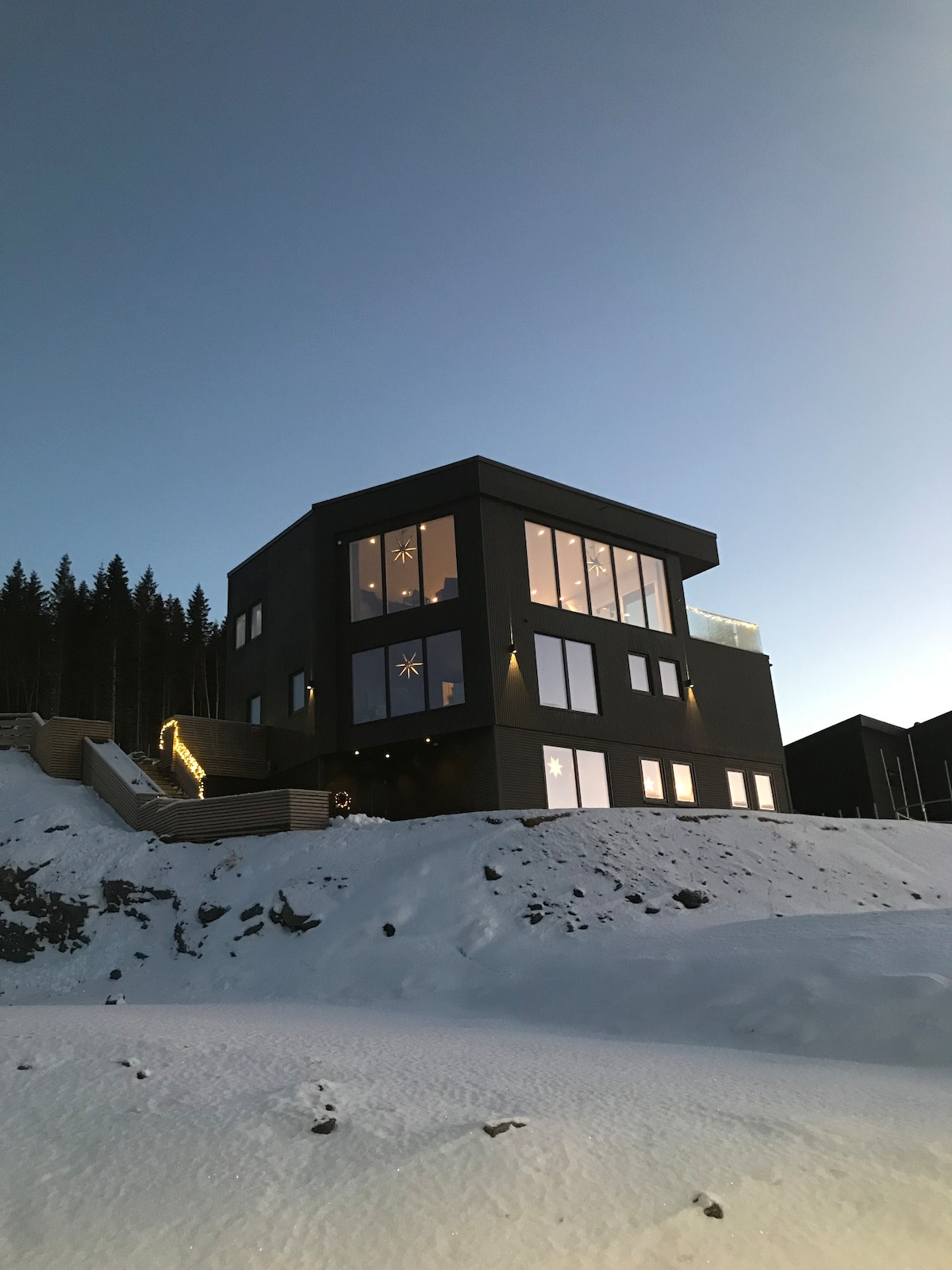 A modern, two-story structure is set against a snowy landscape, featuring large windows that reflect the evening sky. The building is designed with a flat roof and geometric lines, while a pathway illuminated by soft lighting leads to the entrance.