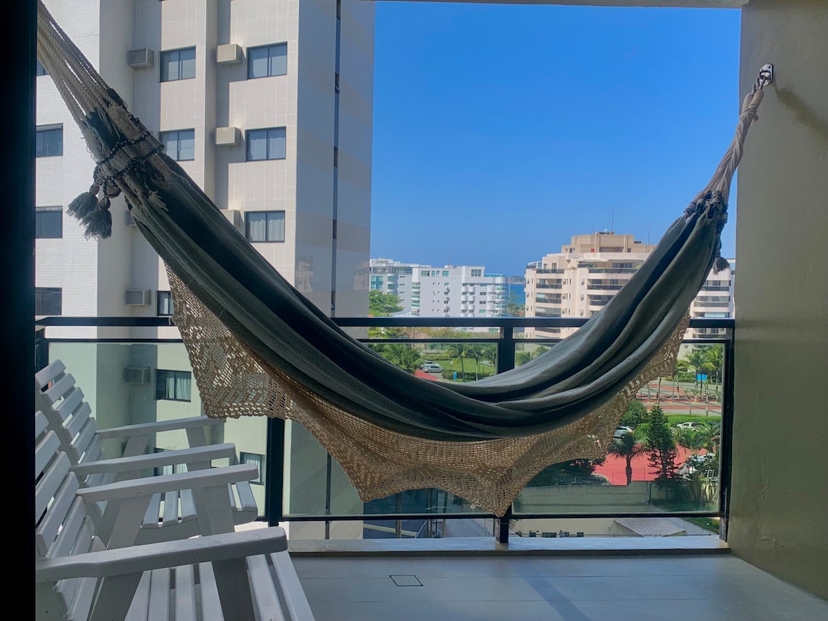 A cozy balcony features a traditional hammock, inviting relaxation. Two white chairs are positioned alongside, with a view of the distant buildings and clear blue sky visible beyond the glass railings.