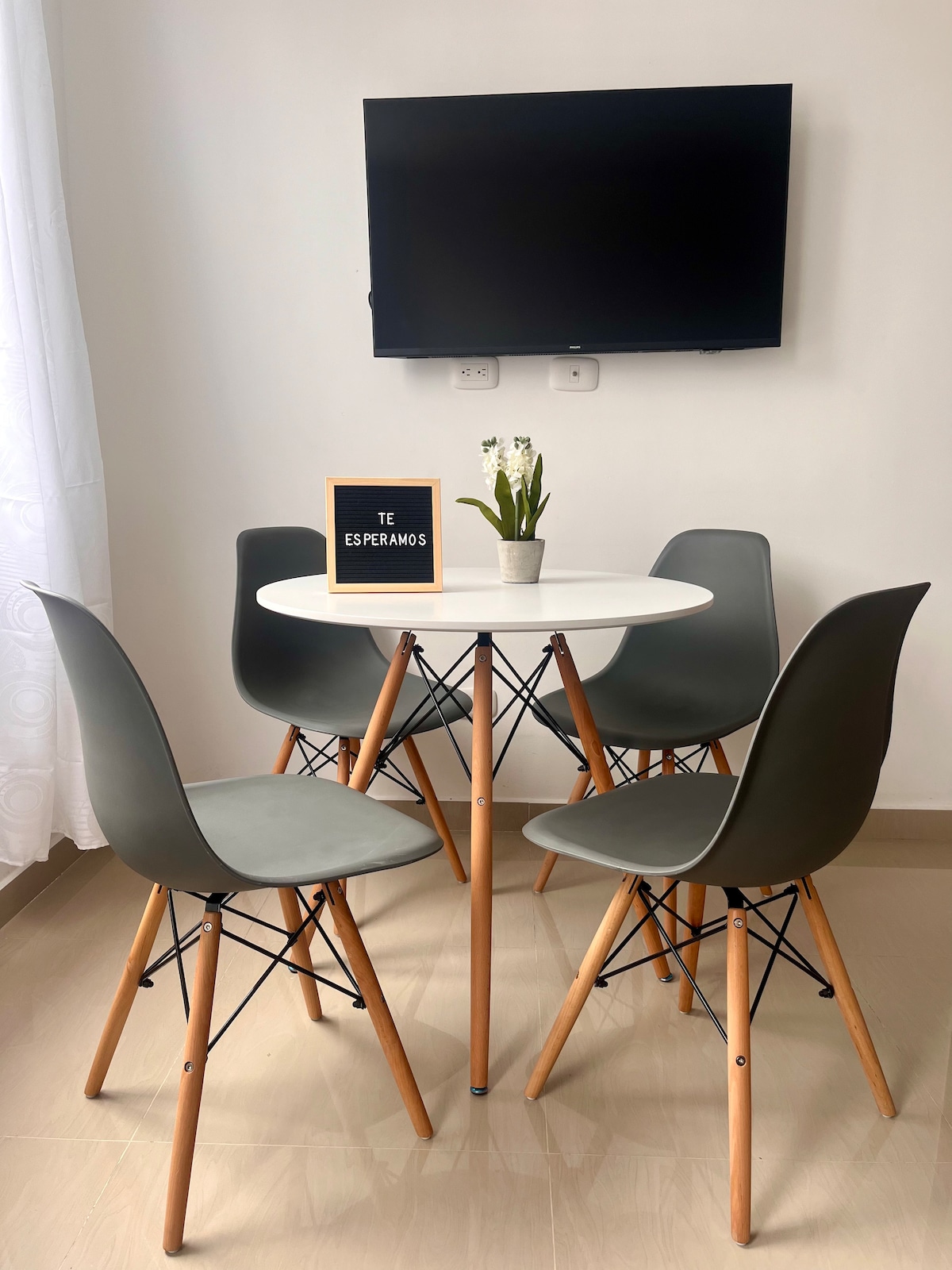 A modern dining area is presented with a circular white table surrounded by four gray chairs featuring wooden legs. A small sign, 'Te Esperamos', is placed on the table, alongside a decorative plant. A wall-mounted TV is visible above the table.