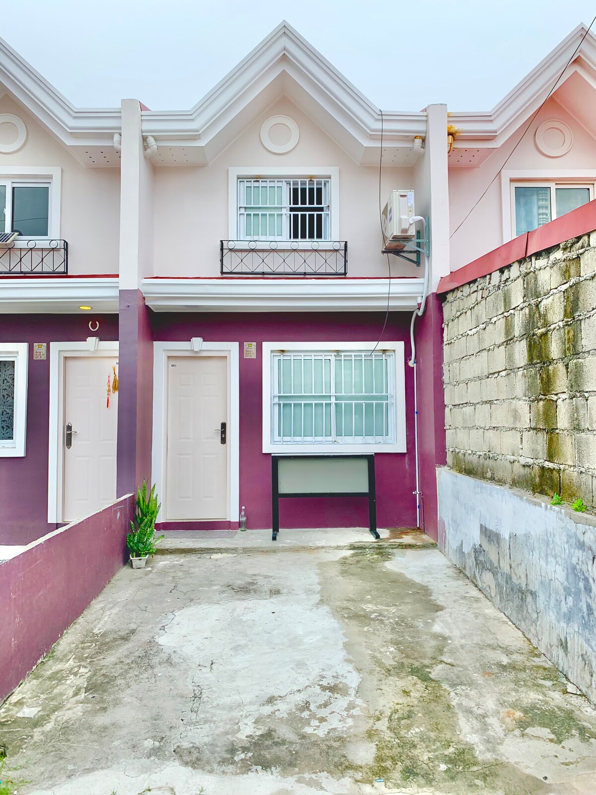 The exterior of the apartment is showcased with a two-tone color scheme. A small pathway leads to the front door, while a designated parking area is visible. The building features a pitched roof and several windows with clear glass panes, providing natural light.