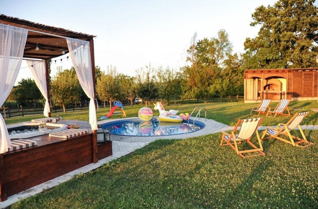 An outdoor area showcases a private swimming pool surrounded by lush greenery. Poolside loungers and a shaded cabana provide relaxation options. Colorful pool floats rest on the water's surface, while a wooden structure offers additional seating and shelter, enhancing the tranquil ambiance.