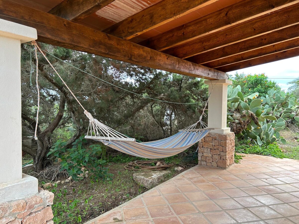 A hammock is gently suspended under a rustic wooden roof, surrounded by lush greenery. The ground is covered with terracotta tiles, and nearby, characteristic plants like cacti can be seen, showcasing the natural ambiance of the outdoor space.