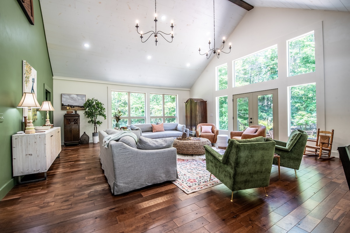 A spacious living area features large windows that invite natural light and offer views of the surrounding greenery. Comfortable seating includes a mix of sofas and chairs arranged around a central coffee table, with wooden flooring adding warmth to the space.