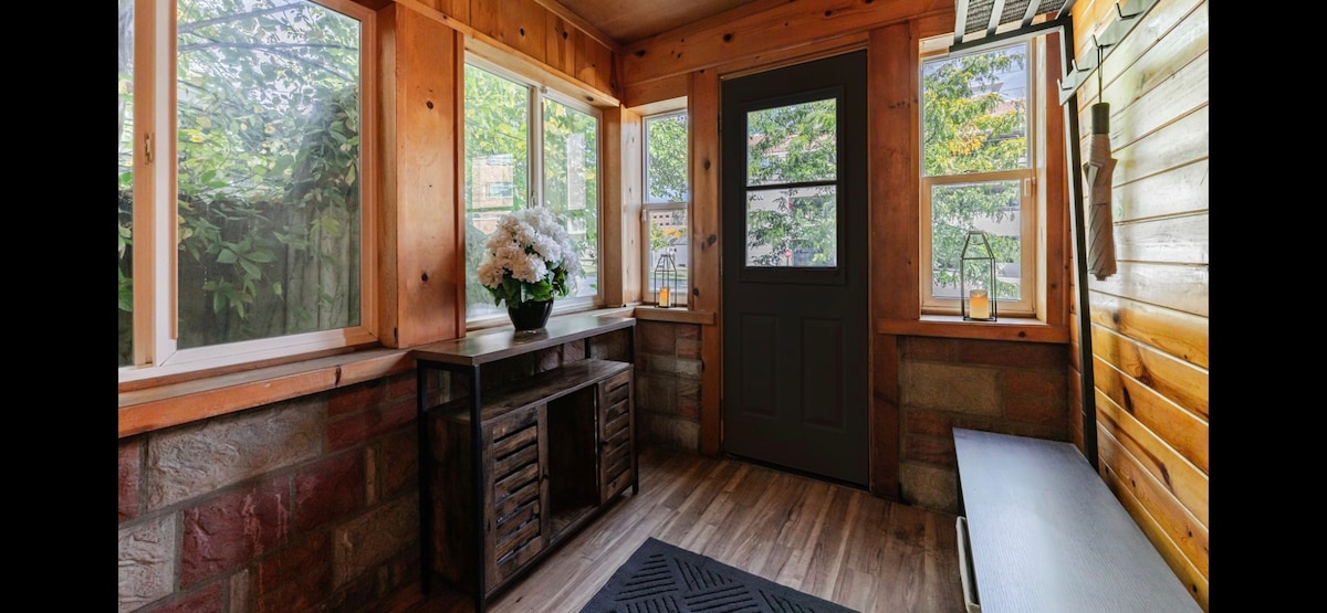 A welcoming entryway features large windows providing natural light and views of greenery. A dark side table with a floral arrangement is positioned against the wall, while a textured area rug lies beneath. The door is framed by light wood paneling, creating a cozy atmosphere.