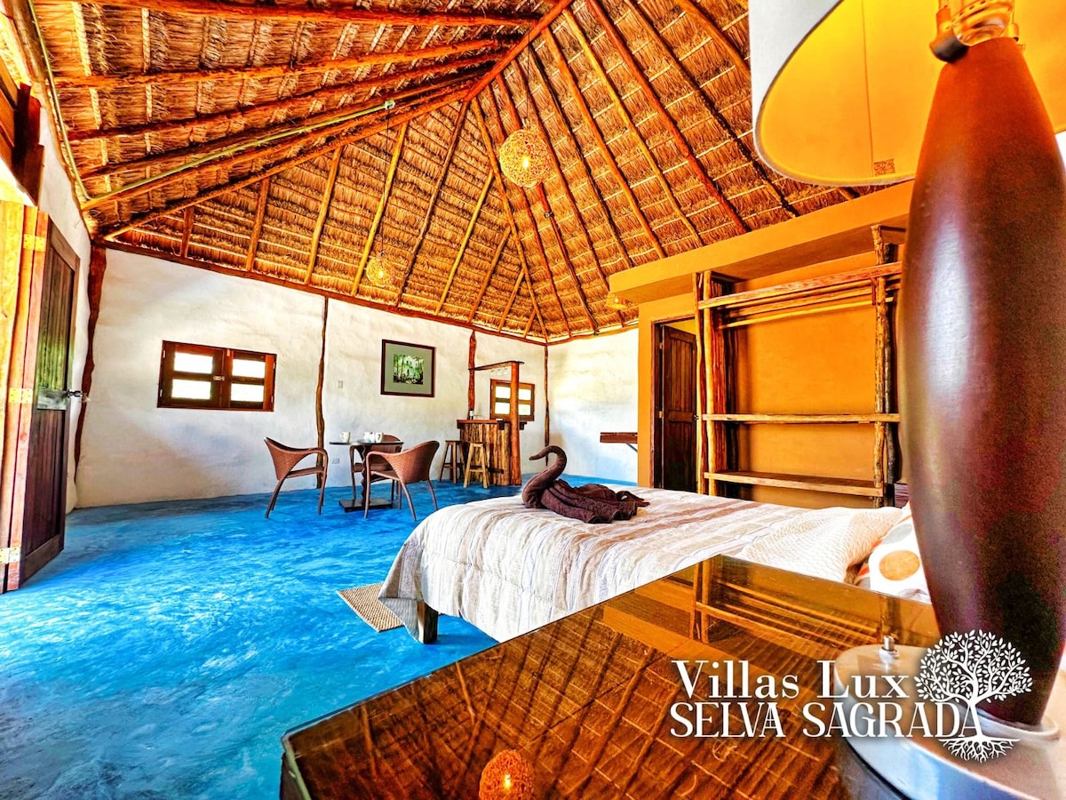 A spacious room features a thatched ceiling and vibrant blue flooring. A queen-sized bed is adorned with a decorative blanket, while natural light filters through wooden windows. A small dining area with two chairs is set in one corner, enhancing the welcoming environment.