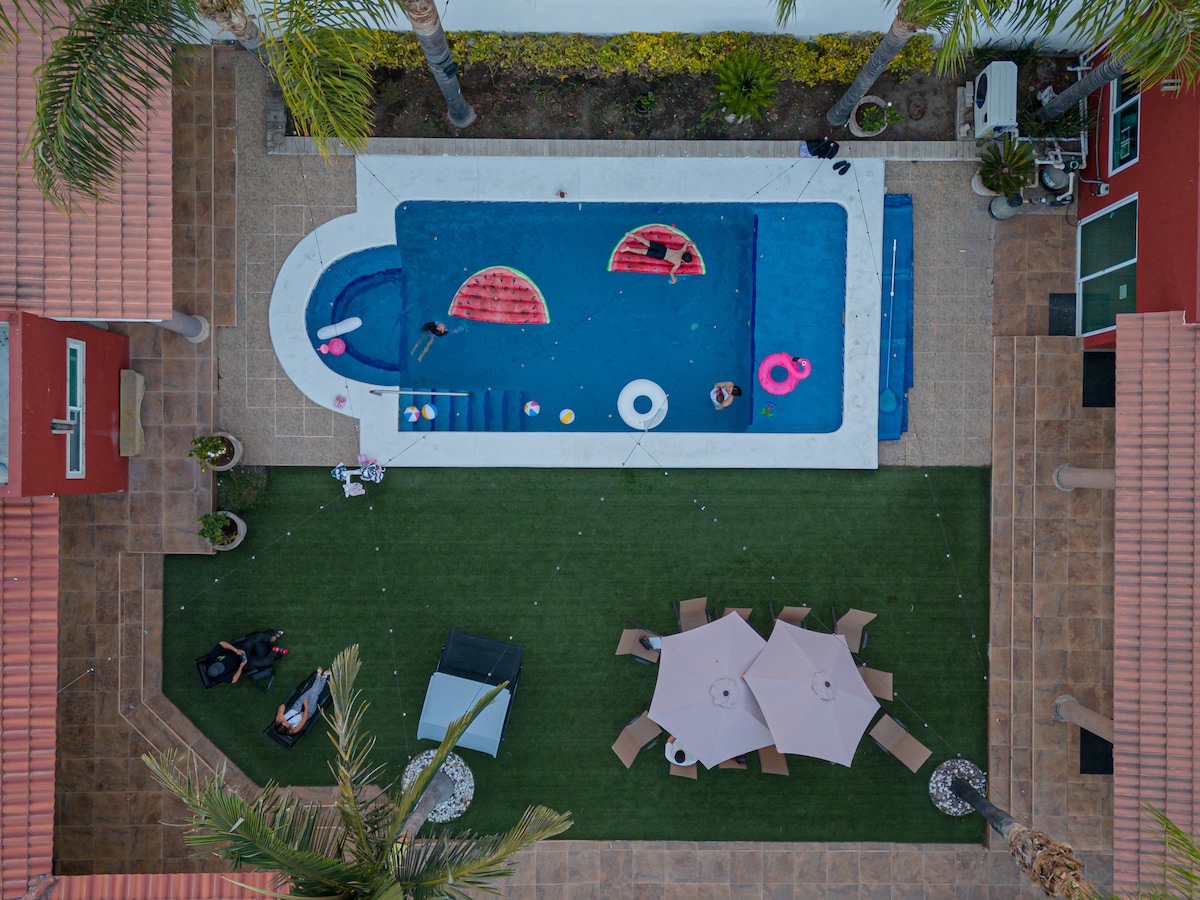 An aerial view showcases a private pool surrounded by a landscaped area. Colorful pool floats rest on the water, and a shaded seating area with a table and chairs is visible on the adjacent lawn. Palm trees frame the scene, enhancing the outdoor space.