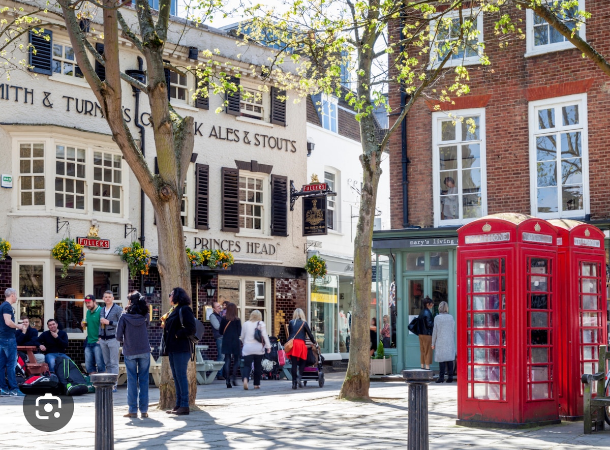 A lively street scene captures people gathering outside local establishments. The iconic red telephone box stands prominently, surrounded by trees with fresh green leaves. Charming storefronts feature traditional signage and flower-filled window boxes, contributing to the inviting atmosphere of the neighborhood.