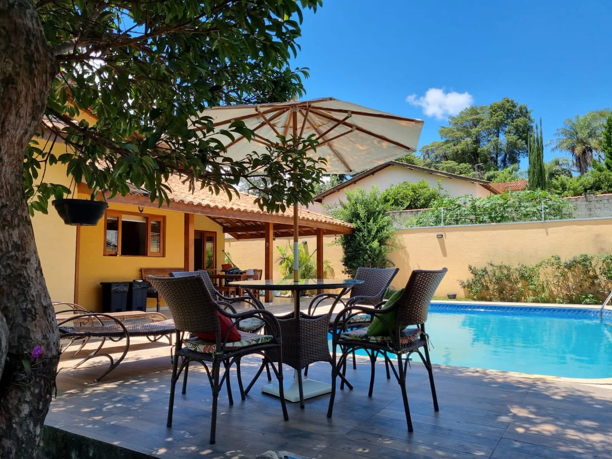 An outdoor seating area is shaded by a large umbrella, featuring a round table surrounded by four wicker chairs. A sparkling pool lies nearby, framed by lush greenery and a sunlit patio, inviting relaxation and social gatherings.