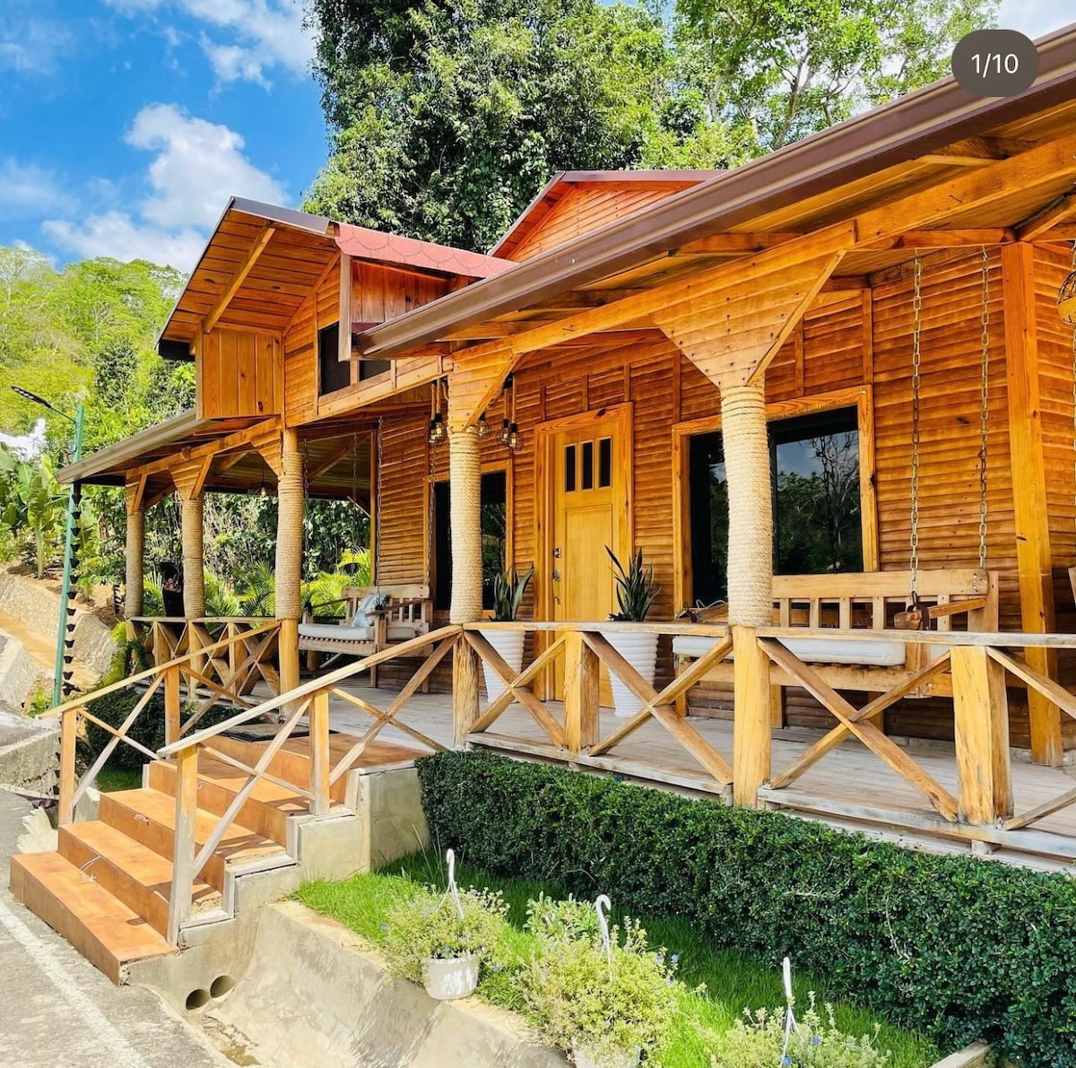 A charming wooden house is featured, showcasing a large porch with ample seating. Sturdy columns support the roof, and stairs lead up to the entryway. Surrounding greenery enhances the serene environment, while large windows allow natural light to illuminate the interior.