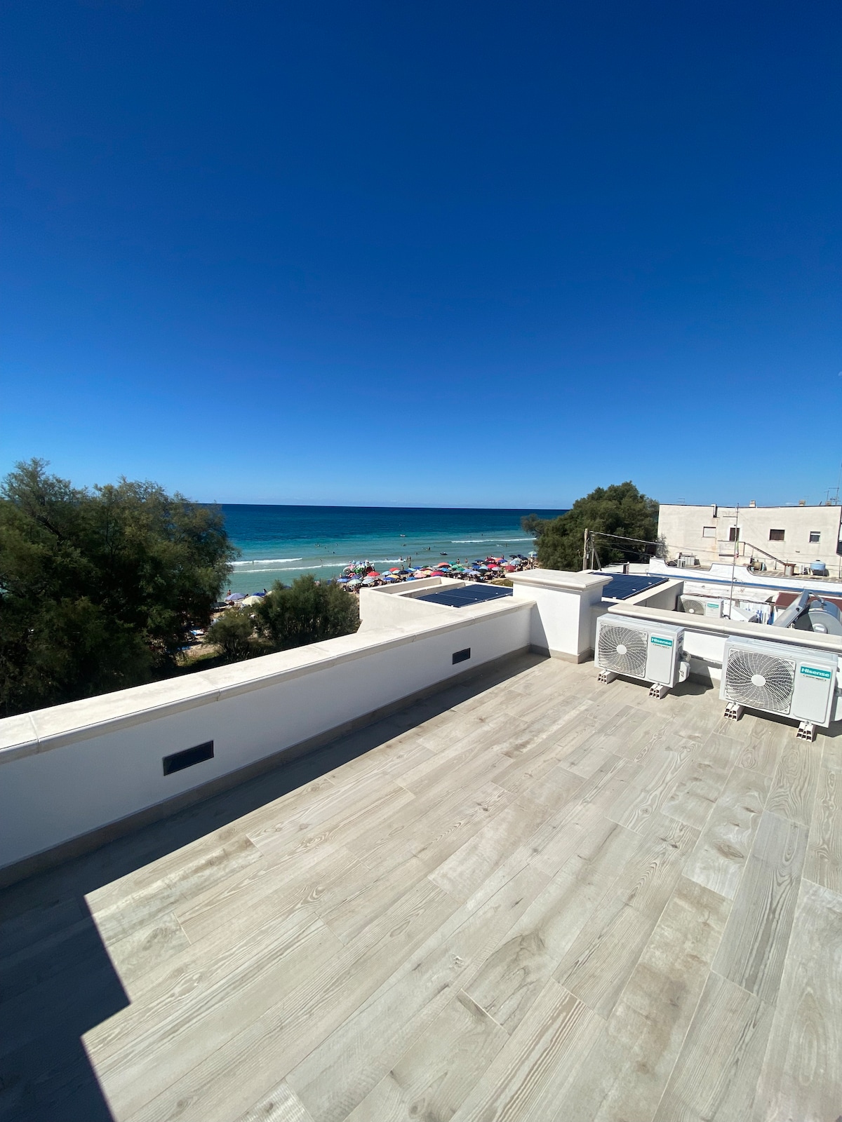A spacious terrace overlooks a sandy beach and the vibrant blue sea. Sunbathers and colorful beach umbrellas are visible below, with air conditioning units positioned discreetly at the edge. Clear skies enhance the panoramic view, creating an open and inviting atmosphere.