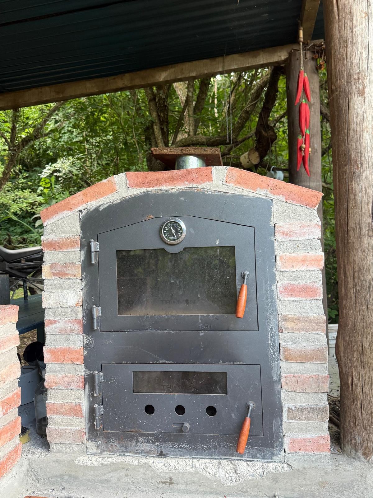 A brick oven is positioned outdoors, featuring a metal door with a handle and a temperature gauge. The structure is surrounded by greenery, creating a rustic ambiance. Tools and cooking supplies are visible nearby, contributing to a communal cooking area.