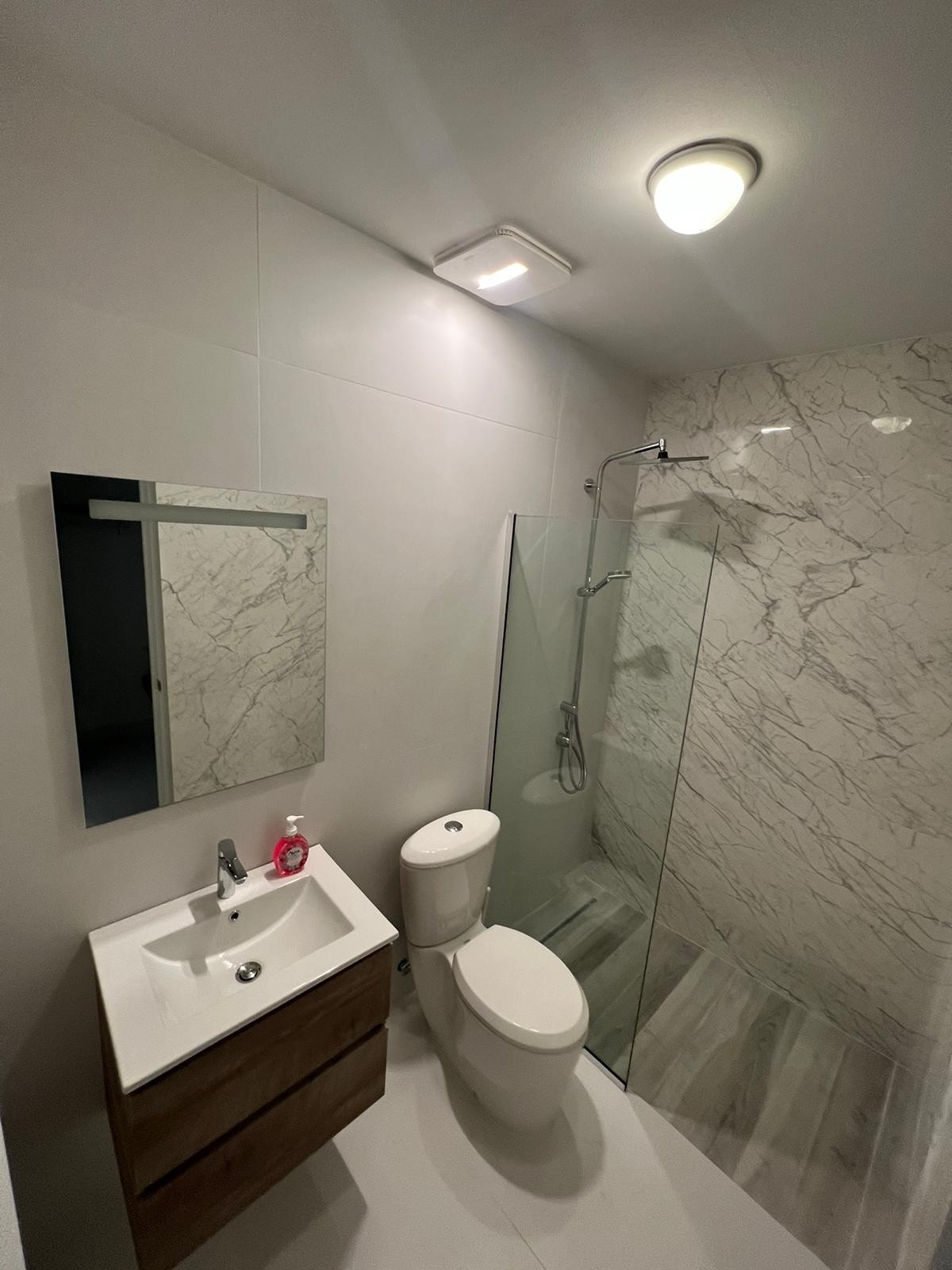 A modern bathroom features light marble walls and a glass-enclosed shower. A sleek vanity with a sink sits below a rectangular mirror, while a toilet is positioned adjacent. Bright overhead lighting illuminates the overall space.