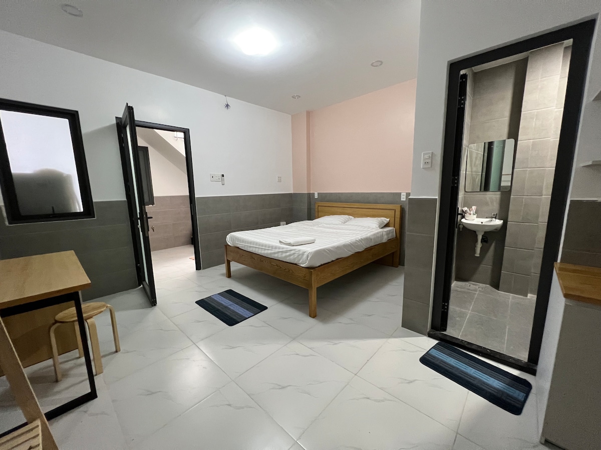 A simple accommodation space features a spacious bed with a light-colored comforter. Two small area rugs provide comfort underfoot. A desk and chair are situated near the entrance, while a compact bathroom is visible with a modern sink and mirror.