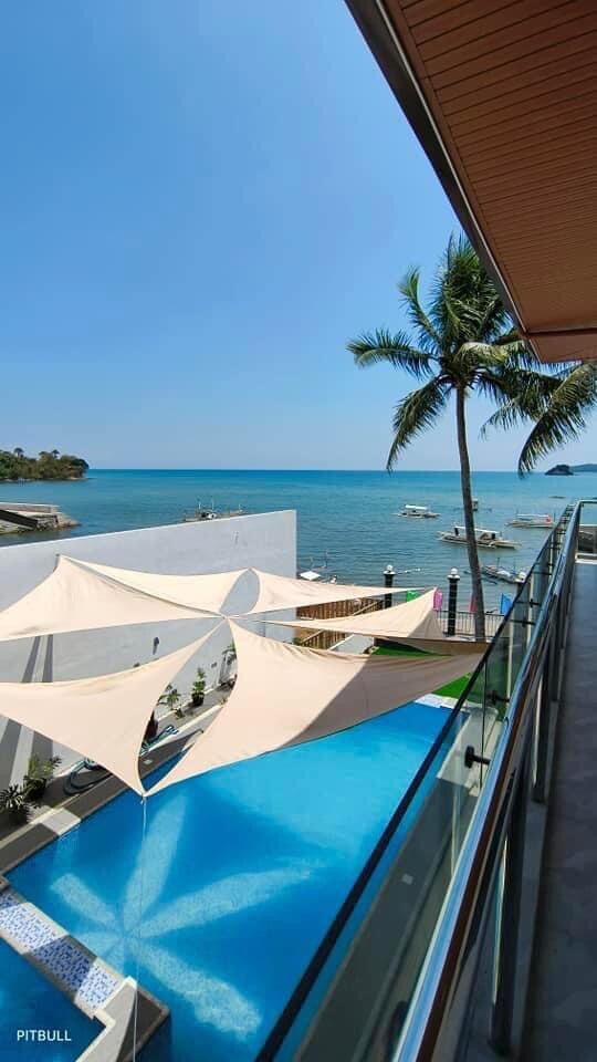 An outdoor view features a refreshing swimming pool bordered by a spacious deck. A series of sail shades provide partial coverage while palm trees sway in the gentle breeze, enhancing the beachside scenery. The tranquil sea and distant boats complete the coastal atmosphere.