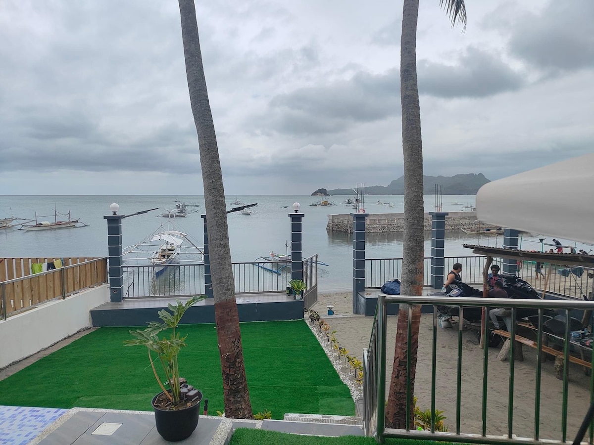 A view of the serene beachfront is captured, showcasing calm waters reflecting a cloudy sky. Palm trees frame the image, while a grassy area and seating spaces are visible in the foreground. Small boats can be seen gently bobbing in the water near the shoreline.