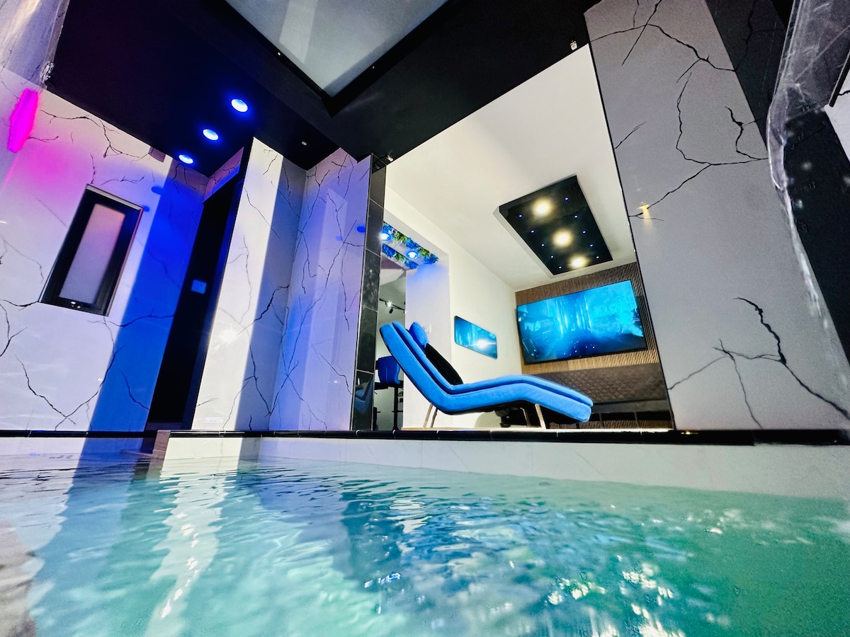 An interior pool is featured, with modern materials and a sleek design. Soft blue and purple lighting create an ambient glow, while a lounge chair is positioned to offer relaxation. The backdrop includes a stylish living area visible through large, reflective surfaces.