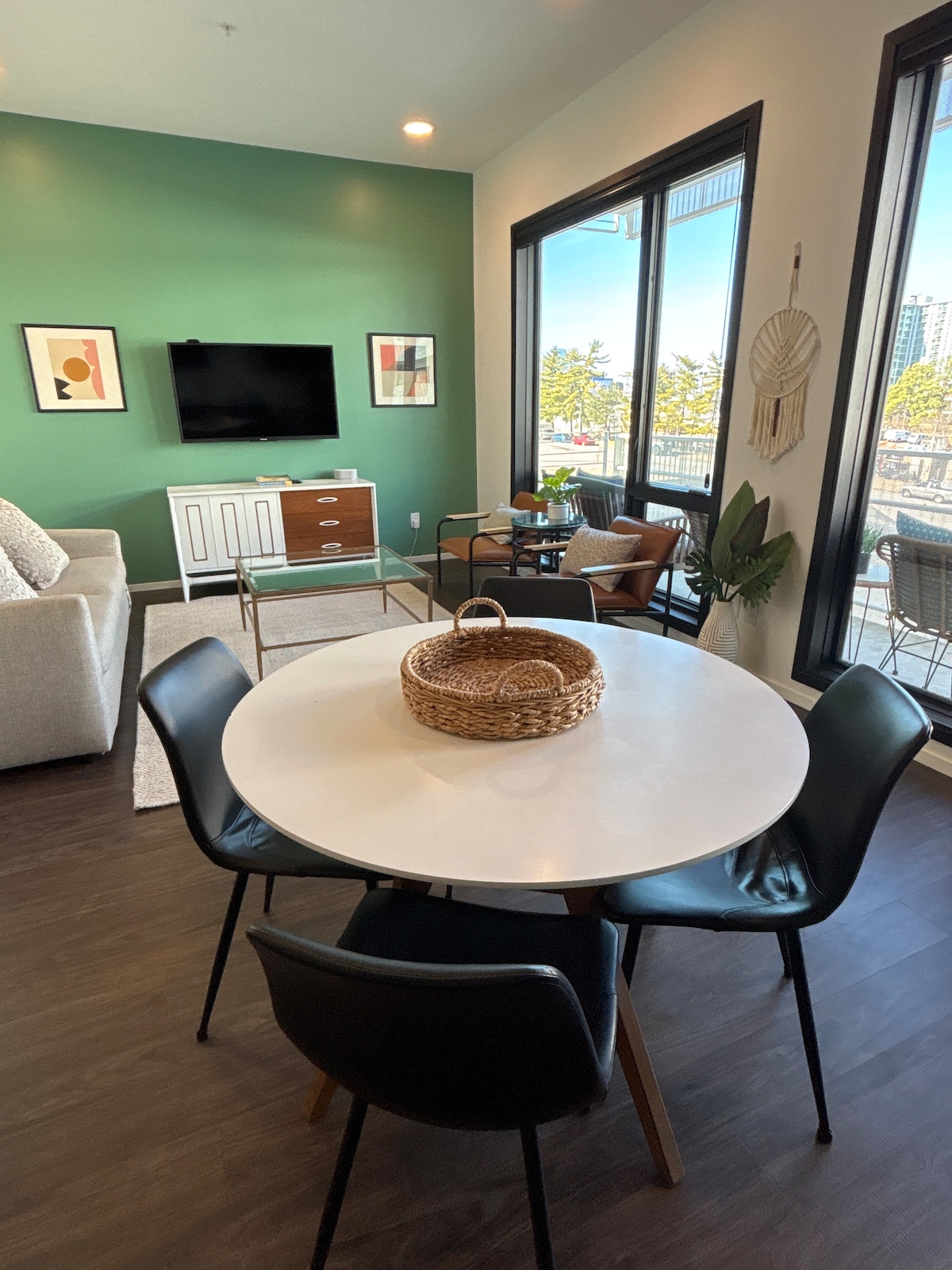 A modern living area features a circular dining table with black chairs surrounded by greenery. A light-colored sofa and a wooden media console with a flat-screen TV are present, and wall-to-ceiling windows offer natural light and views of the balcony.