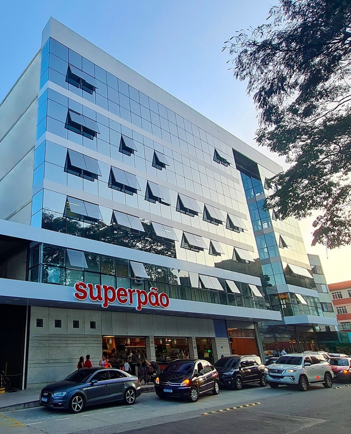 A modern building features a sleek glass facade that reflects the surrounding environment. The entrance showcases the Superpão bakery sign, with patrons gathered outside. Several cars are parked along the street, enhancing the urban atmosphere of the area.