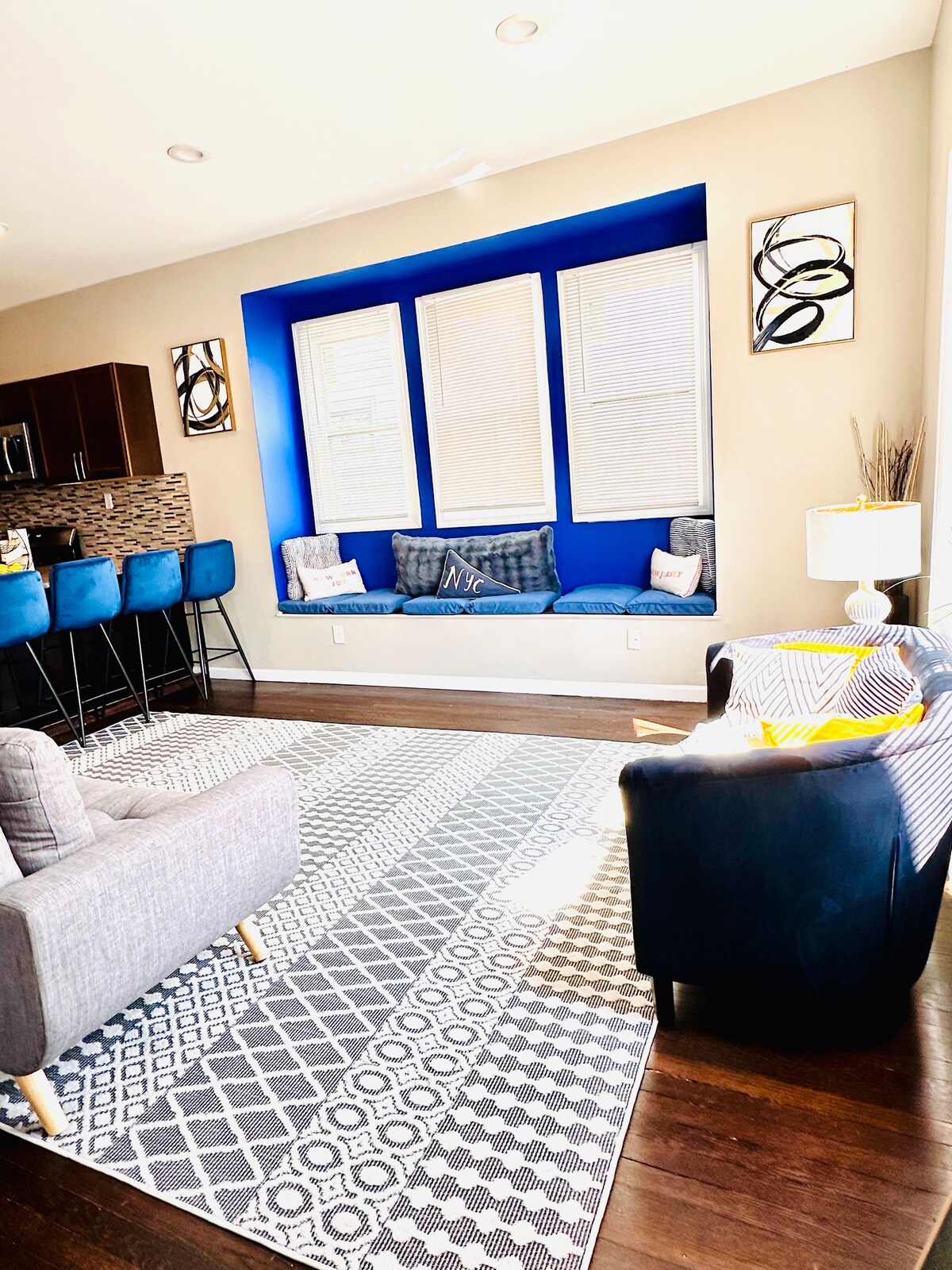 A bright and spacious living area features a cozy seating arrangement with a gray sofa and a blue accent chair. A built-in window seat is adorned with decorative pillows, while natural light streams in through the three windows. Stylish bar stools complement the kitchen area in the background.
