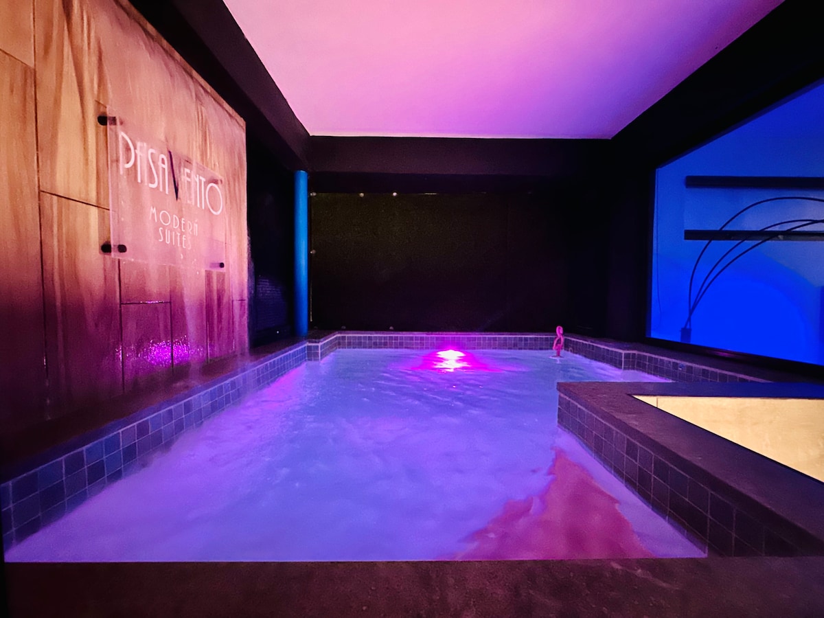 An indoor pool area features calm waters illuminated by colorful lighting, creating a serene ambiance. The walls are designed with modern textures, and the gentle glow reflects off the surface, enhancing the tranquil setting.
